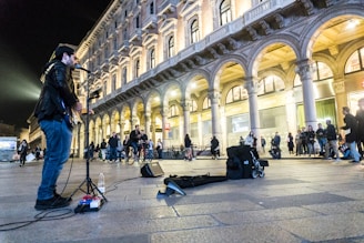 A musician is performing on a city street at night. He is playing an electric guitar in front of a historical building with arches and illuminated windows. Several people stand around watching the performance, while some pass by or sit nearby. The scene has a lively and engaging atmosphere.