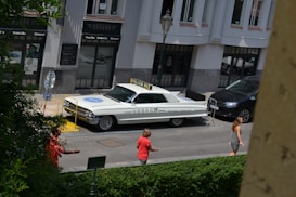 A vintage white car with signage on top and writing on the side is parked on a city street near a building with large windows and 'Hangar' signs. There are a few pedestrians on the sidewalk, one wearing a red shirt and another in a gray outfit. A street lamp is in the background, and there is greenery in the foreground.