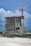 A construction site features a partially built, multi-story concrete building with an attached tower crane. The structure is surrounded by construction materials and equipment. The background shows a cloudy sky with some blue patches.