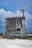 A construction site features a partially built, multi-story concrete building with an attached tower crane. The structure is surrounded by construction materials and equipment. The background shows a cloudy sky with some blue patches.