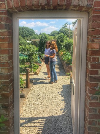 A garden scene visible through a brick archway, featuring two people conversing on a gravel path bordered by lush green plants and trees under a clear blue sky.