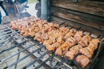 Several skewers of seasoned and grilled meat are placed over a barbecue grill with glowing charcoal beneath. The meat appears to be well-cooked with a slightly charred exterior, suggesting it is kebab or shashlik. The setting includes a wooden backdrop, and there's a hint of smoke rising from the grill, indicating active cooking.