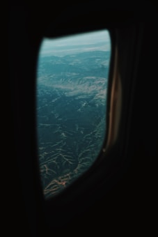 The image captures a scenic view of mountainous terrain as seen through an airplane window. The landscape features rugged mountain ranges with subtle variations in elevation, hinting at valleys and peaks. The sky is visible in the background, with a hazy atmosphere that suggests high altitude and distant horizons.