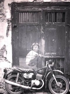 A black and white photograph depicts a motorcycle parked in front of an old wooden door with a painted mural of a child peering from behind, creating a striking juxtaposition of art and reality. The door is weathered with visible cracks and peeling paint which adds to the vintage atmosphere.