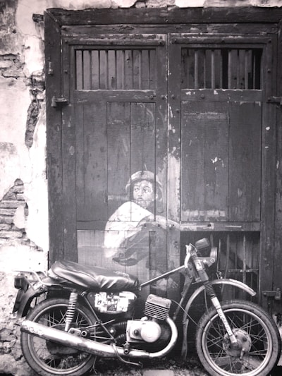 A black and white photograph depicts a motorcycle parked in front of an old wooden door with a painted mural of a child peering from behind, creating a striking juxtaposition of art and reality. The door is weathered with visible cracks and peeling paint which adds to the vintage atmosphere.