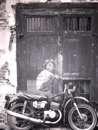 A black and white photograph depicts a motorcycle parked in front of an old wooden door with a painted mural of a child peering from behind, creating a striking juxtaposition of art and reality. The door is weathered with visible cracks and peeling paint which adds to the vintage atmosphere.