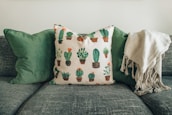 A comfortable sofa adorned with two green cushions and a decorative pillow featuring various cactus designs. A soft, beige throw is draped over the right side of the sofa.