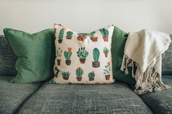 A comfortable sofa adorned with two green cushions and a decorative pillow featuring various cactus designs. A soft, beige throw is draped over the right side of the s