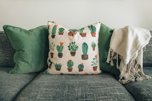 A comfortable sofa adorned with two green cushions and a decorative pillow featuring various cactus designs. A soft, beige throw is draped over the right side of the sofa.