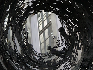 Several bronze sculptures appear to be reaching outwards, surrounded by swirling, jagged metallic elements. These figures seem to be caught in a vortex of dark metal, set against a backdrop of a modern building with large windows.