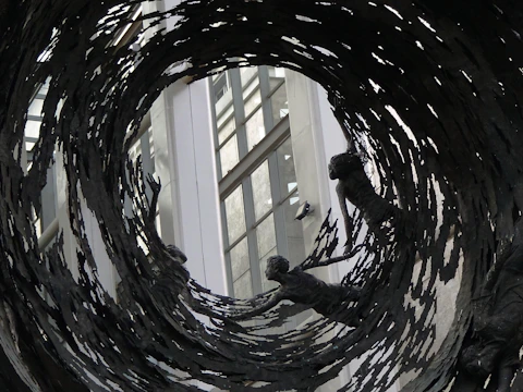 Several bronze sculptures appear to be reaching outwards, surrounded by swirling, jagged metallic elements. These figures seem to be caught in a vortex of dark metal, set against a backdrop of a modern building with large windows.