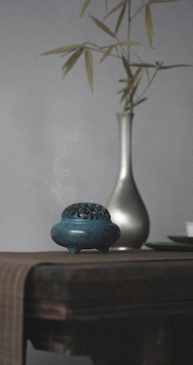 A dimly lit scene featuring a ceramic incense burner with smoke rising delicately from it. The burner is placed on a wooden table draped with a textured fabric. In the background, a slender metallic vase holds a few bamboo stalks, adding an element of minimalistic decor.