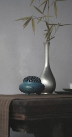 A dimly lit scene featuring a ceramic incense burner with smoke rising delicately from it. The burner is placed on a wooden table draped with a textured fabric. In the background, a slender metallic vase holds a few bamboo stalks, adding an element of minimalistic decor.