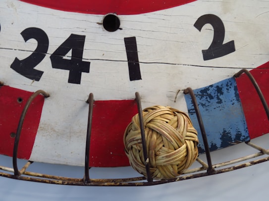 A close-up view of a traditional wooden board painted in red, white, and blue with large black numbers (24, 1, 2) displayed. A small wicker ball is placed within a metal cage attached to the board.