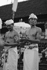 Two men dressed in traditional attire, possibly for a cultural or religious ceremony. They are bare-chested and wear cloths around their waists and headpieces. One man is holding a shell-like object. The background suggests an outdoor setting, possibly with a traditional building and various ceremonial decorations.