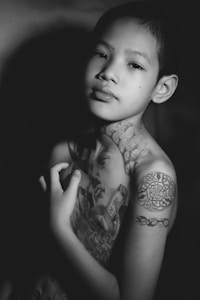 A young person is featured with intricate tattoos covering their torso and shoulder, appearing in a black and white photograph. The tattoos include detailed designs and portrait-like elements. The subject has a contemplative expression and is partially illuminated, creating a play of light and shadow.