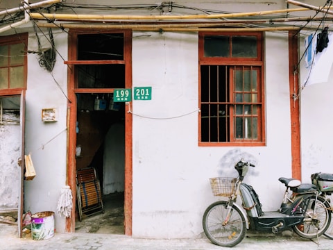 A weathered building facade with two wooden-framed windows, one of which is open. A doorway leading inside the building displays hanging items. A bicycle with a basket on the front is parked against the wall. Electrical wires and pipes run above the windows and door. A bucket with cleaning supplies sits near the entrance.