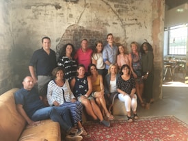 A group of people are gathered in a rustic, industrial-style room with exposed brick walls and worn plaster. They are posing for a photo on a brown leather sofa and a patterned rug. The atmosphere is casual and relaxed.