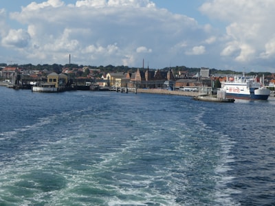 A coastal town with a view of a port and distinctive historic buildings along the waterfront. The sea in the foreground has visible waves from a passing boat. A ferry labeled 'HH-Ferries' is docked at the pier. The sky is partly cloudy, creating a picturesque and lively atmosphere.