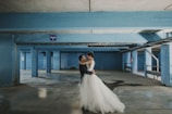A couple is embracing in a spacious parking garage with blue walls and columns. The bride is wearing a white gown, and the groom is in a dark suit, creating a contrast against the industrial setting. Dim lighting and shadows accentuate the cement floor and the various textures of the environment.