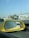 A close-up view of a yellow car's side mirror on the left, reflecting a blurry image of traffic behind. The mirror has Arabic text printed on it. In the background, there is a bright blue sky and several vehicles, including a white truck and some cars.