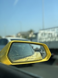 A close-up view of a yellow car's side mirror on the left, reflecting a blurry image of traffic behind. The mirror has Arabic text printed on it. In the background, there is a bright blue sky and several vehicles, including a white truck and some cars.