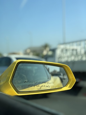 A close-up view of a yellow car's side mirror on the left, reflecting a blurry image of traffic behind. The mirror has Arabic text printed on it. In the background, there is a bright blue sky and several vehicles, including a white truck and some cars.