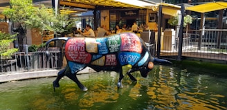 A colorful bull sculpture stands in a shallow pool of water. The bull is painted with different labeled cuts of meat, such as rib, brisket, and chuck. The background shows a lively outdoor restaurant with people dining under yellow awnings, surrounded by greenery.