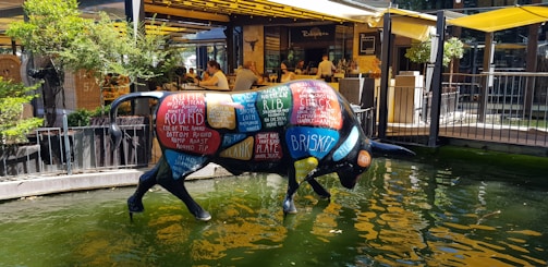 A colorful bull sculpture stands in a shallow pool of water. The bull is painted with different labeled cuts of meat, such as rib, brisket, and chuck. The background shows a lively outdoor restaurant with people dining under yellow awnings, surrounded by greenery.