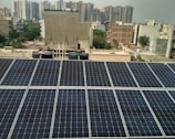 Rows of solar panels are installed on a rooftop in an urban setting, with various residential and commercial buildings in the background. The panels are arranged in neat rows, reflecting sunlight. In the distance, high-rise buildings contrast with the smaller structures in the foreground. Water tanks and pipes are visible alongside the panels.