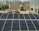 Rows of solar panels are installed on a rooftop in an urban setting, with various residential and commercial buildings in the background. The panels are arranged in neat rows, reflecting sunlight. In the distance, high-rise buildings contrast with the smaller structures in the foreground. Water tanks and pipes are visible alongside the panels.