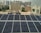 Rows of solar panels are installed on a rooftop in an urban setting, with various residential and commercial buildings in the background. The panels are arranged in neat rows, reflecting sunlight. In the distance, high-rise buildings contrast with the smaller structures in the foreground. Water tanks and pipes are visible alongside the panels.