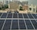 Rows of solar panels are installed on a rooftop in an urban setting, with various residential and commercial buildings in the background. The panels are arranged in neat rows, reflecting sunlight. In the distance, high-rise buildings contrast with the smaller structures in the foreground. Water tanks and pipes are visible alongside the panels.