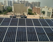 Rows of solar panels are installed on a rooftop in an urban setting, with various residential and commercial buildings in the background. The panels are arranged in neat rows, reflecting sunlight. In the distance, high-rise buildings contrast with the smaller structures in the foreground. Water tanks and pipes are visible alongside the panels.