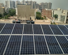 Rows of solar panels are installed on a rooftop in an urban setting, with various residential and commercial buildings in the background. The panels are arranged in neat rows, reflecting sunlight. In the distance, high-rise buildings contrast with the smaller structures in the foreground. Water tanks and pipes are visible alongside the panels.