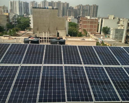 Rows of solar panels are installed on a rooftop in an urban setting, with various residential and commercial buildings in the background. The panels are arranged in neat rows, reflecting sunlight. In the distance, high-rise buildings contrast with the smaller structures in the foreground. Water tanks and pipes are visible alongside the panels.
