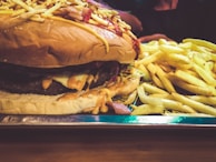 A large cheeseburger topped with crunchy ingredients like potato sticks, served alongside a generous portion of golden French fries. The burger is layered with melted cheese and sauces, sitting on a shiny metal tray.