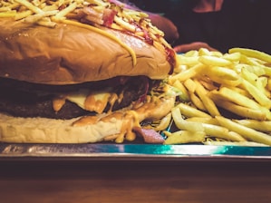A large cheeseburger topped with crunchy ingredients like potato sticks, served alongside a generous portion of golden French fries. The burger is layered with melted cheese and sauces, sitting on a shiny metal tray.
