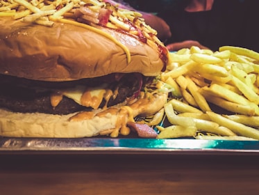 A large cheeseburger topped with crunchy ingredients like potato sticks, served alongside a generous portion of golden French fries. The burger is layered with melted cheese and sauces, sitting on a shiny metal tray.