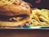 A large cheeseburger topped with crunchy ingredients like potato sticks, served alongside a generous portion of golden French fries. The burger is layered with melted cheese and sauces, sitting on a shiny metal tray.