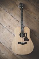 An acoustic guitar is lying flat on a wooden floor with a natural finish. The guitar has a light wood body with a dark pickguard and a visible rosette around the soundhole.