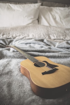 A wooden acoustic guitar is placed on a soft, textured gray blanket. The background shows pillows with white pillowcases, suggesting a cozy and relaxed setting, possibly a bedroom.