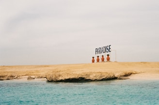 A sandy beach with a rocky formation meets a calm, turquoise ocean. Large potted plants line the edge of the beach next to a sign that reads 'PARADISE'. The sky is overcast, creating a serene and peaceful atmosphere.