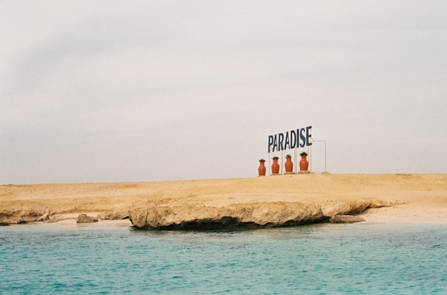 A sandy beach with a rocky formation meets a calm, turquoise ocean. Large potted plants line the edge of the beach next to a sign that reads 'PARADISE'. The sky is overcast, creating a serene and peaceful atmosphere.