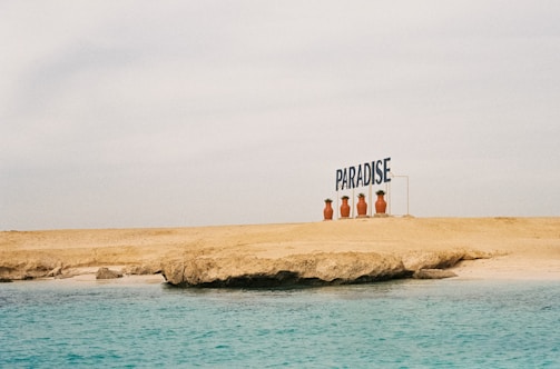 A sandy beach with a rocky formation meets a calm, turquoise ocean. Large potted plants line the edge of the beach next to a sign that reads 'PARADISE'. The sky is overcast, creating a serene and peaceful atmosphere.