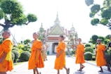 A group of young boys dressed in bright orange robes walking in front of a temple with intricate details and statues of mythical figures. Lush greenery and meticulously trimmed bushes surround the scene. The ornate temple structure features elaborate decorative elements.