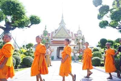 A group of young boys dressed in bright orange robes walking in front of a temple with intricate details and statues of mythical figures. Lush greenery and meticulously trimmed bushes surround the scene. The ornate temple structure features elaborate decorative elements.