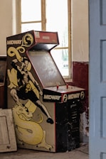 A dusty, vintage Midway arcade machine with a fighting game theme is placed against a wall in an old room. The machine's artwork features a martial artist in a fighting stance, accompanied by a yellow dragon. The room has peeling paint, large windows letting in natural light, and some old furniture scattered around.
