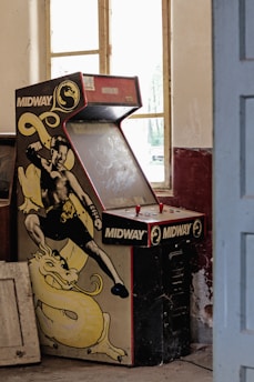 A dusty, vintage Midway arcade machine with a fighting game theme is placed against a wall in an old room. The machine's artwork features a martial artist in a fighting stance, accompanied by a yellow dragon. The room has peeling paint, large windows letting in natural light, and some old furniture scattered around.