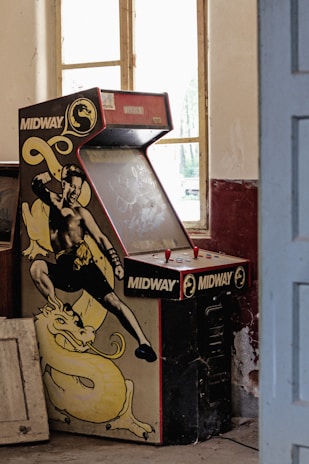 A dusty, vintage Midway arcade machine with a fighting game theme is placed against a wall in an old room. The machine's artwork features a martial artist in a fighting stance, accompanied by a yellow dragon. The room has peeling paint, large windows letting in natural light, and some old furniture scattered around.